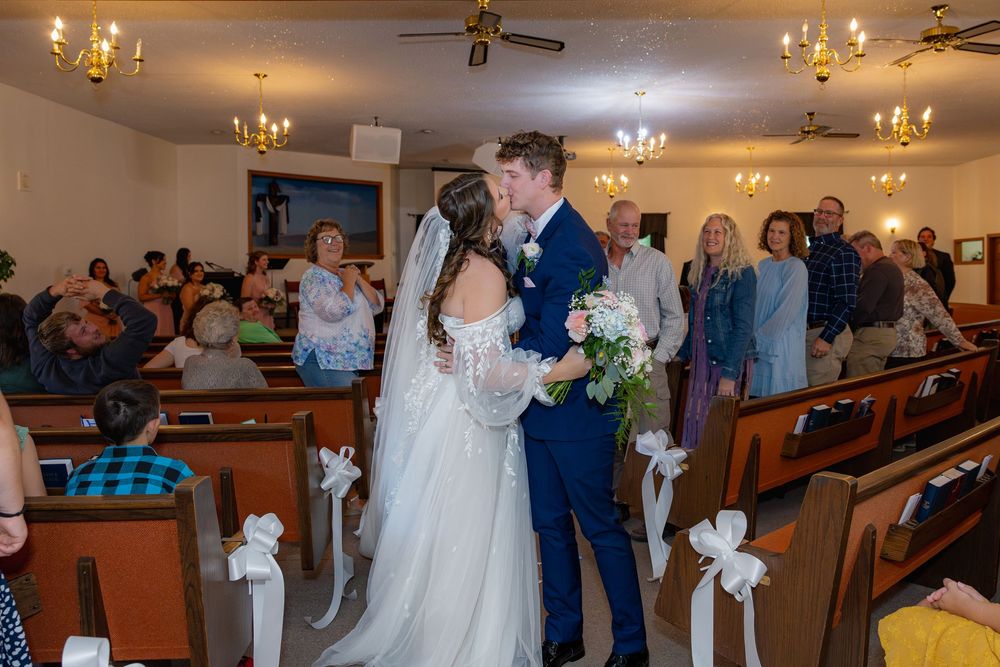 Wedding ceremony venue with wooden pews, white bows, and warm chandelier lighting.