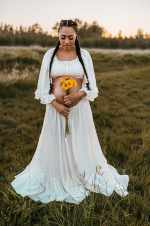 A person in a flowing white dress holds vibrant yellow sunflowers in a golden-lit meadow at sunset.