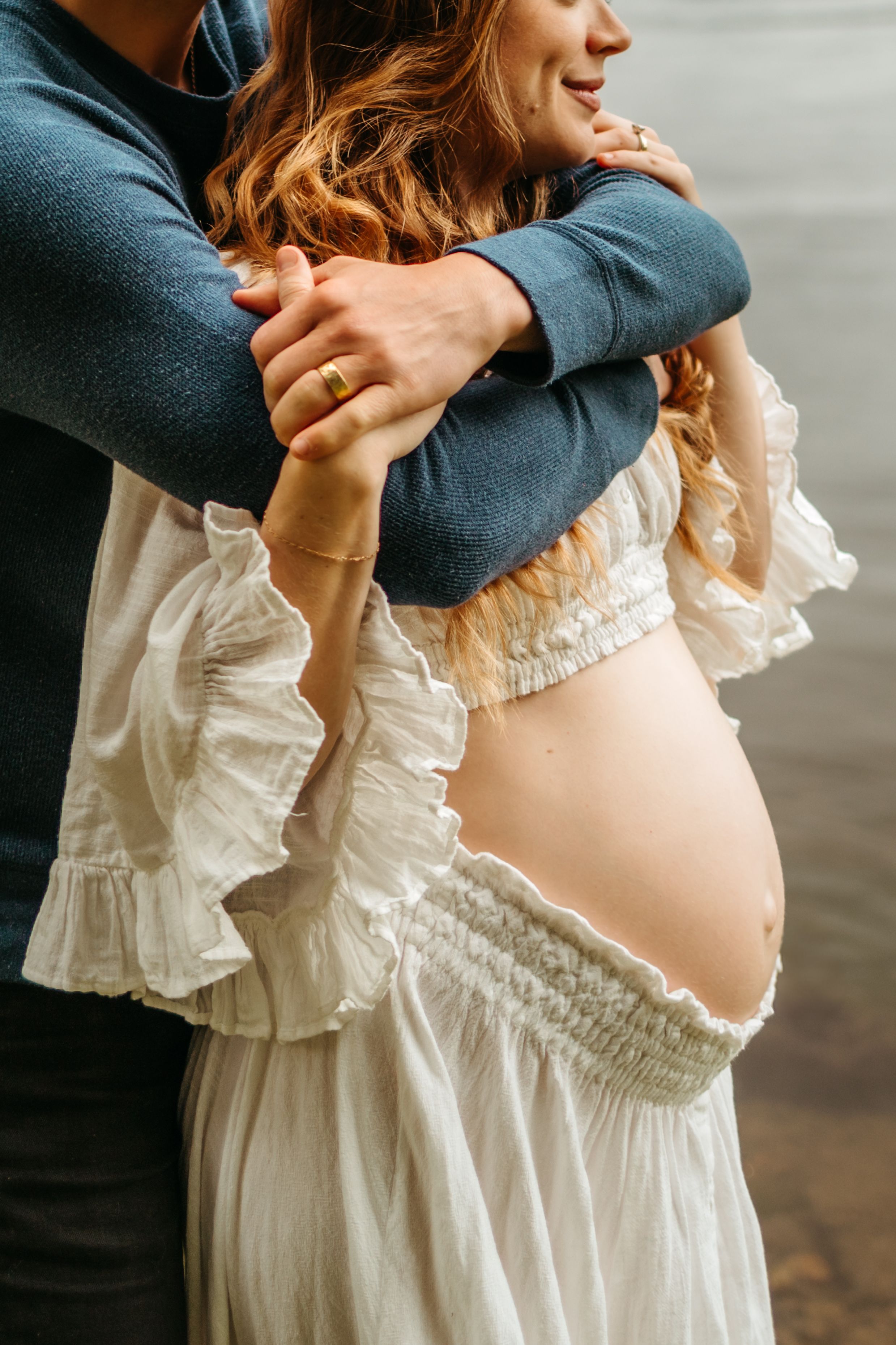 An intimate maternity portrait shows a tender embrace in white lace dress against a moody background.