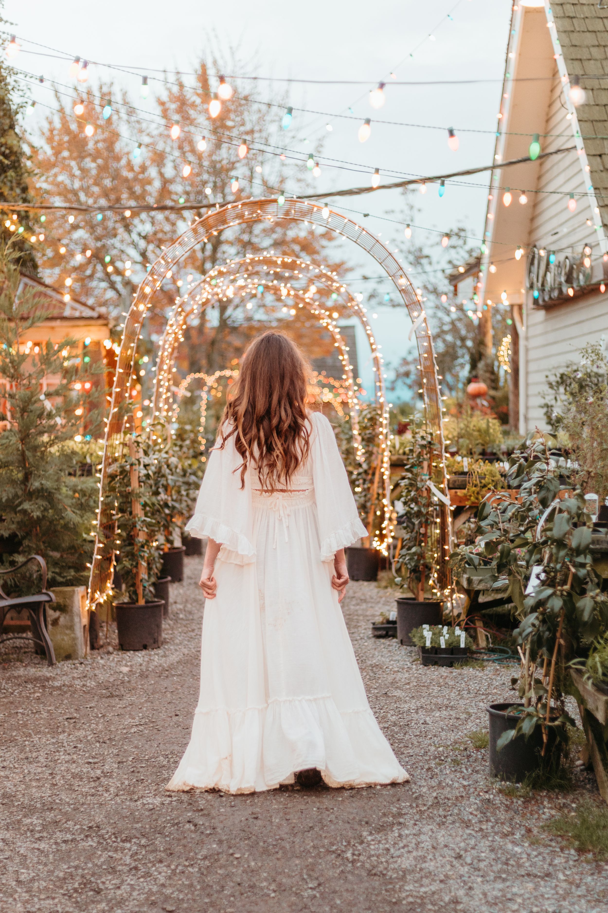 A person in a flowing white wedding dress twirls in a garden decorated with twinkling string lights at dusk.