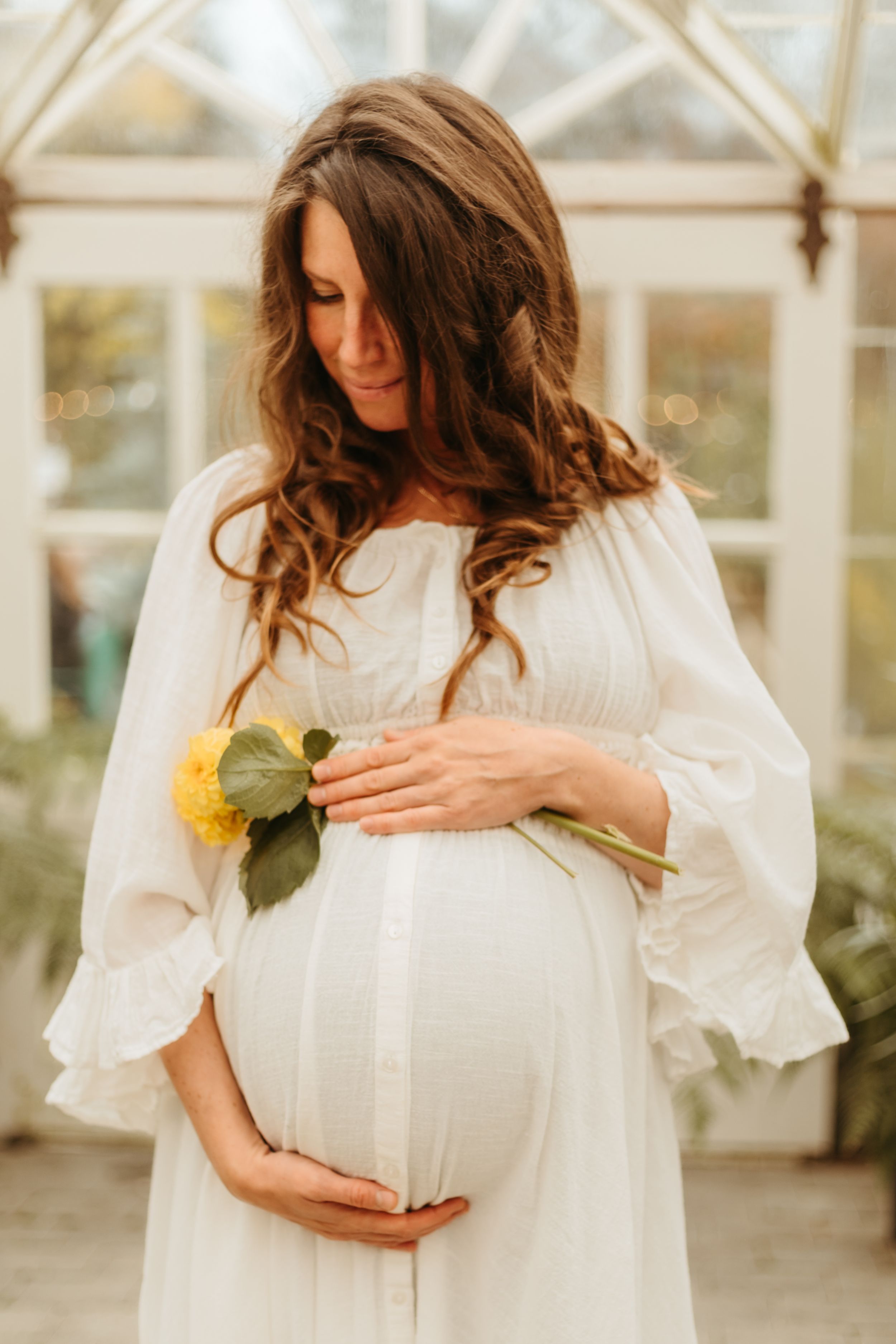 Expectant mother in white flowing dress holds yellow flower in sunlit greenhouse setting.