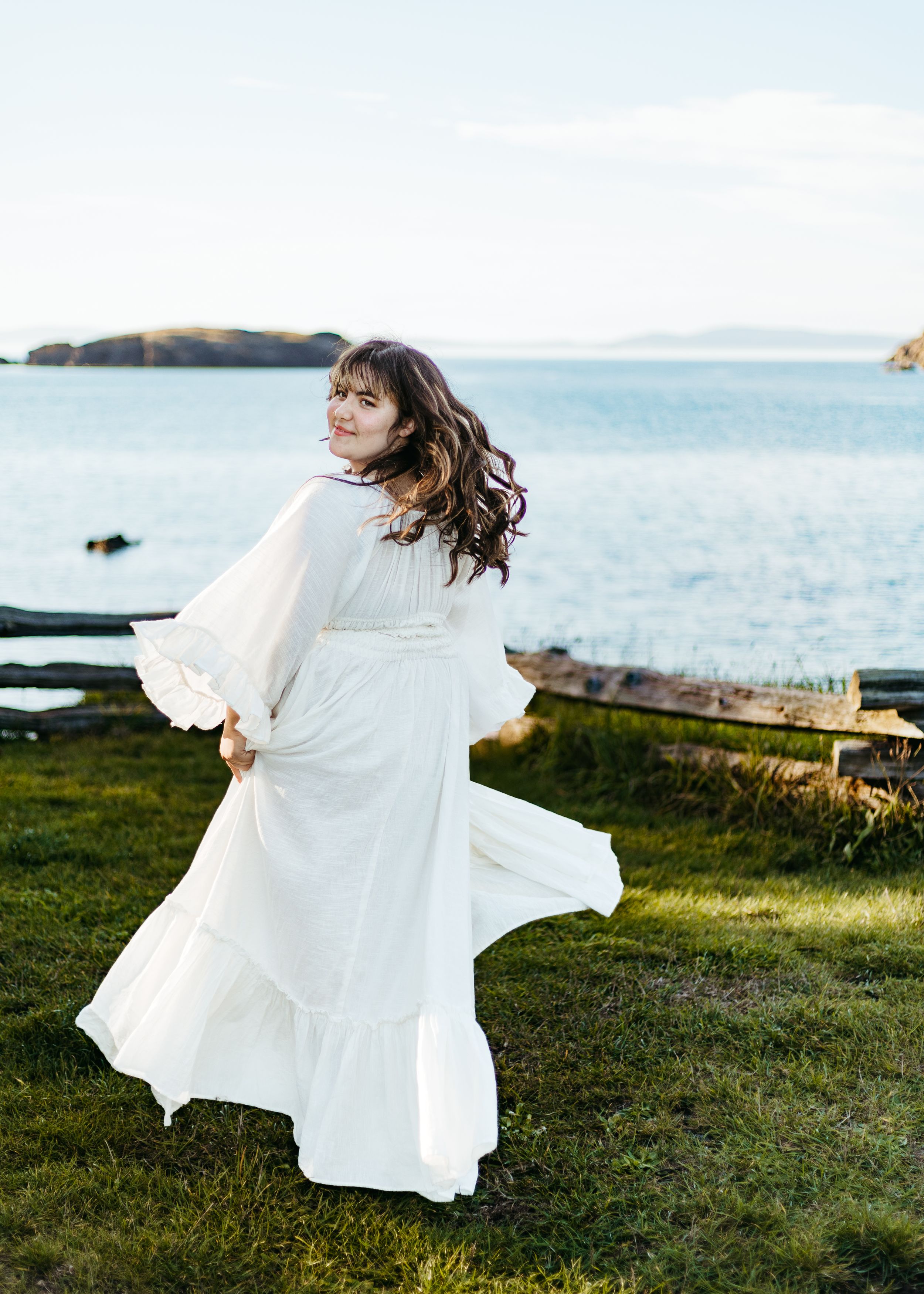 A person in a flowing white dress spins gracefully on a grassy cliff overlooking the ocean at sunset.
