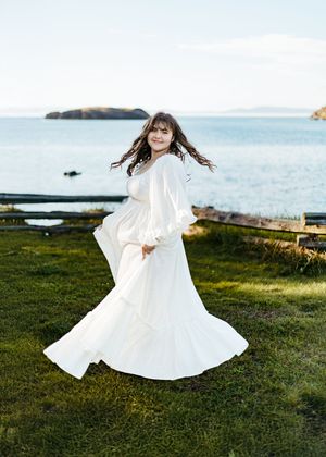 A person in a flowing white dress twirls gracefully on a grassy coastal cliff overlooking the ocean at sunset.