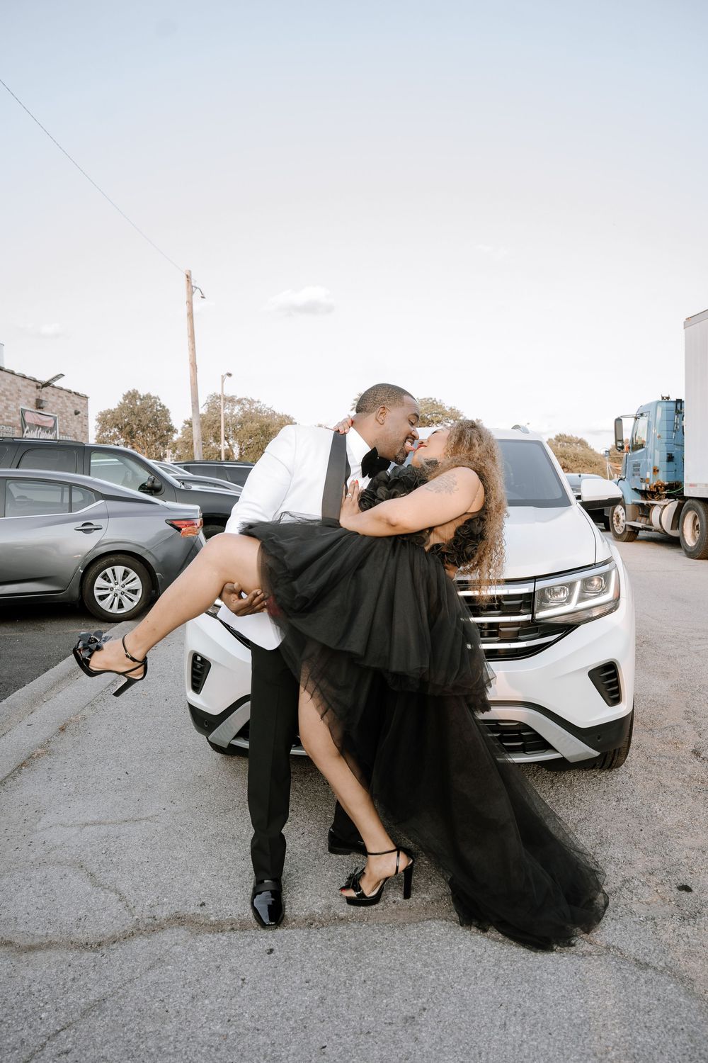 A couple in black attire performs a dramatic dance dip pose in a parking lot with cars in the background.