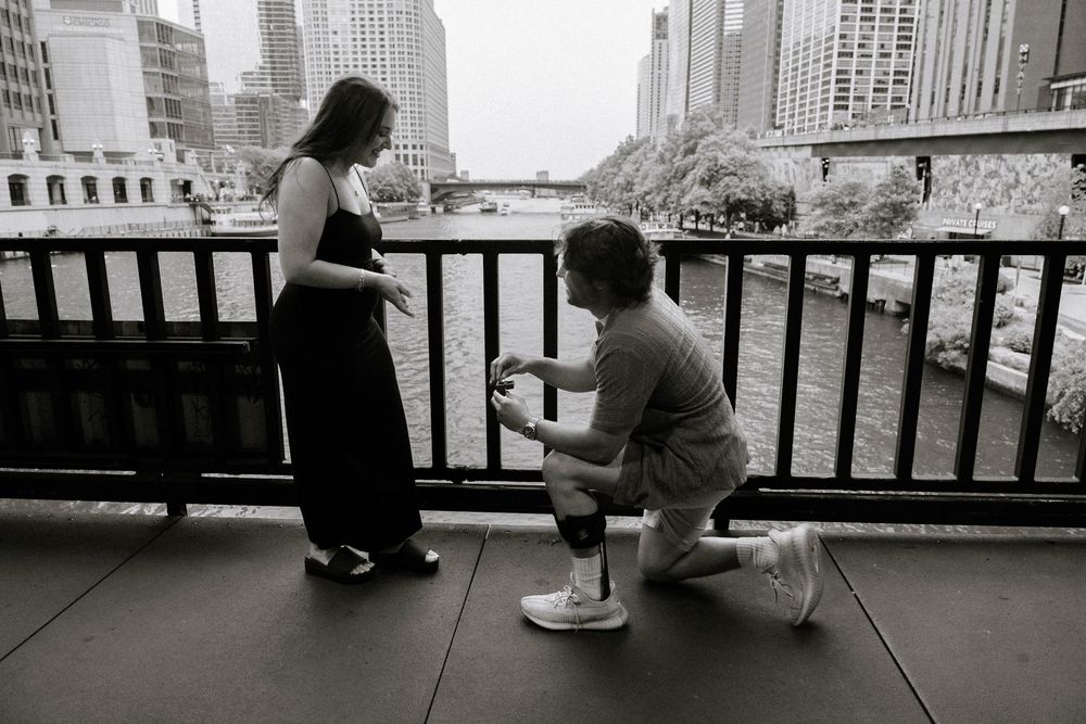 A romantic proposal moment captured in black and white on an urban bridge overlooking a city skyline.