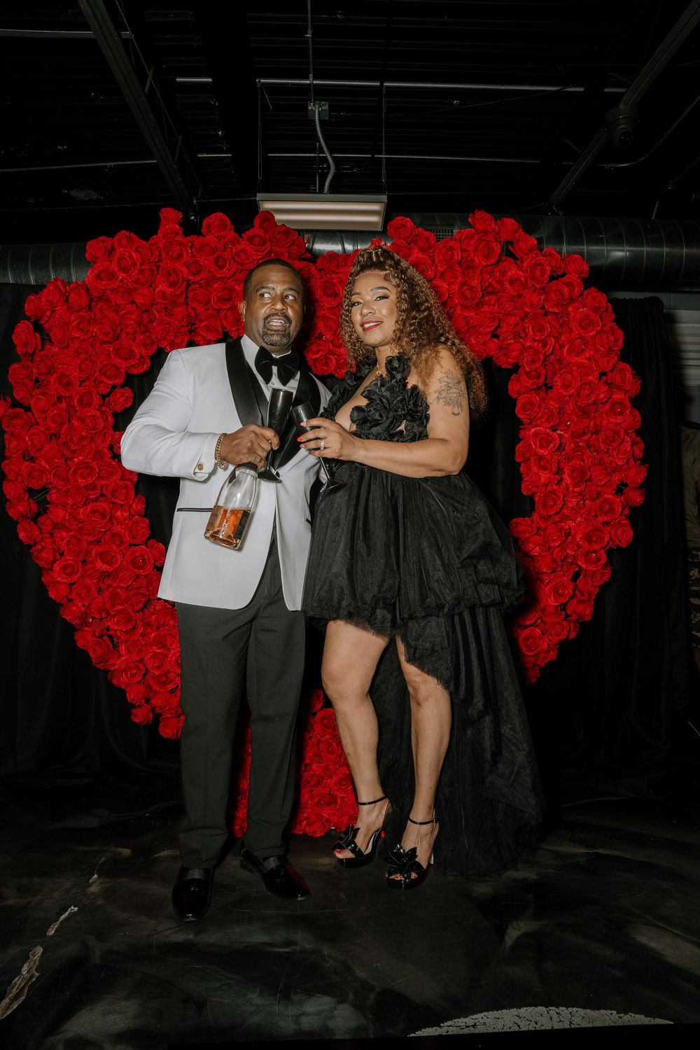 A couple poses together in formal attire against a large red rose heart backdrop at an elegant event.