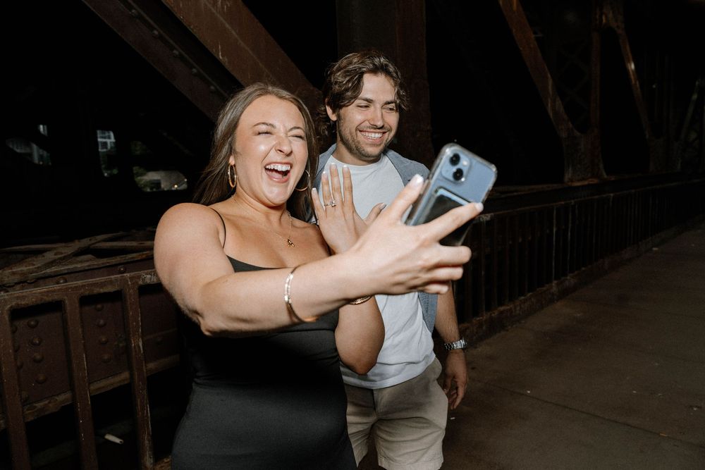A couple in wedding attire takes selfies and laughs together in a dimly lit venue.