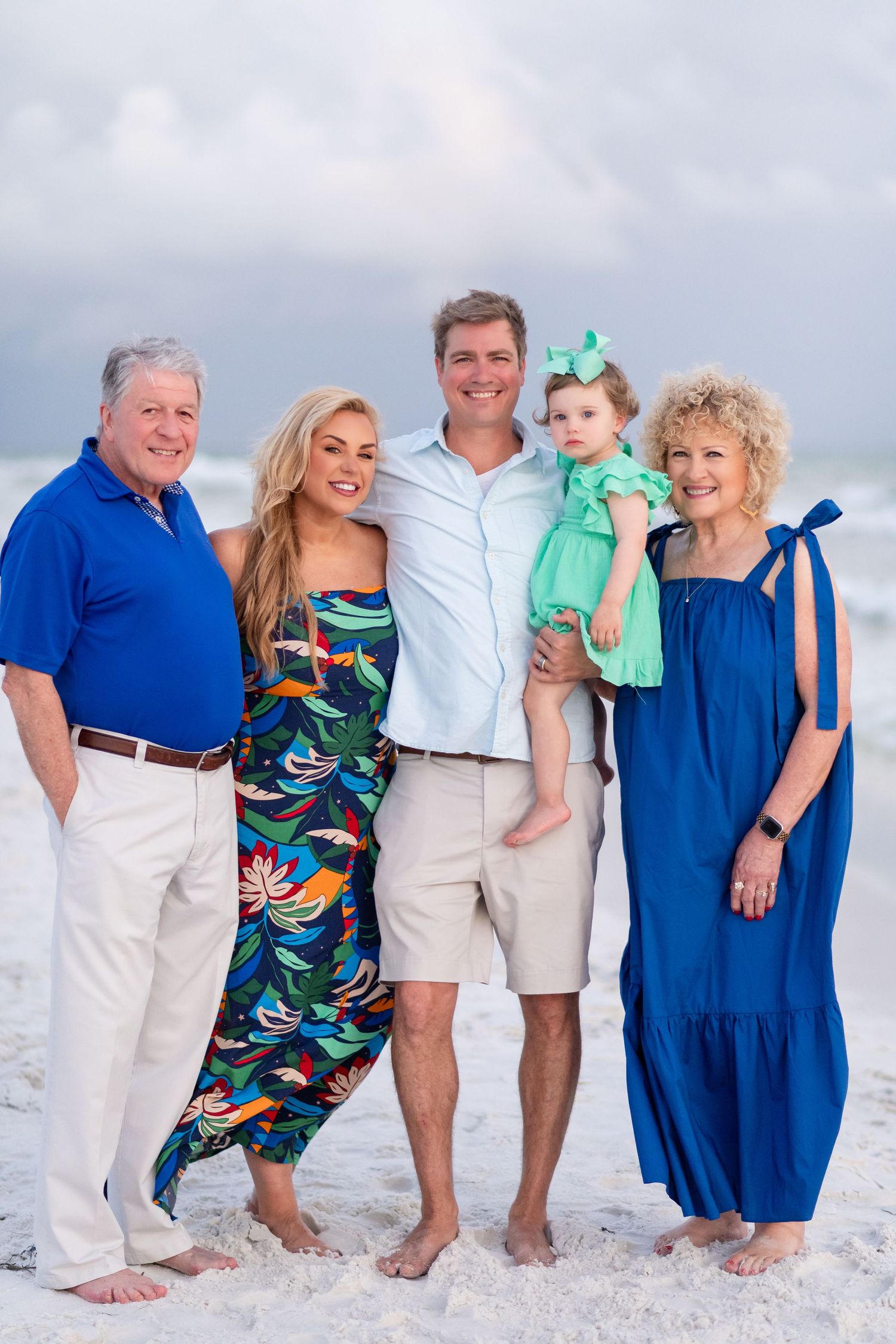 A family of five poses together on a white sandy beach wearing coordinated blue and tropical print outfits.
