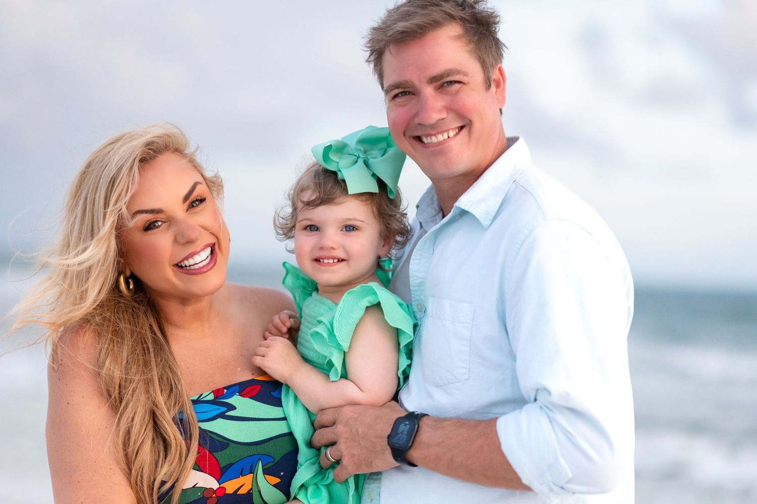 A happy family of three smiles brightly during their beach photoshoot in coordinating outfits.