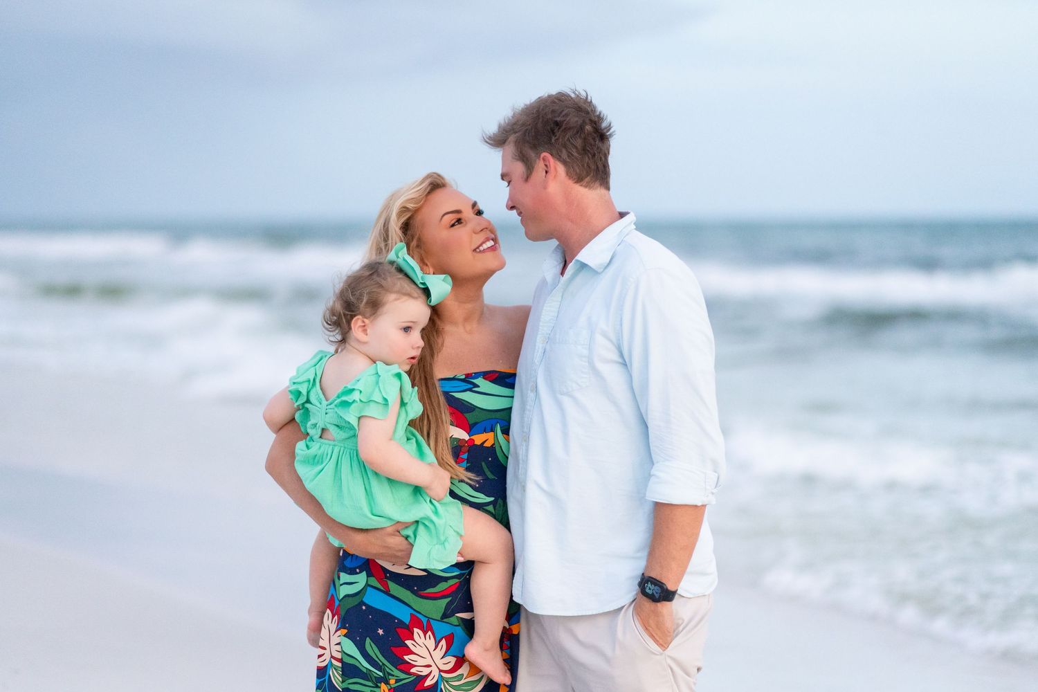 A family embraces while posing for a portrait on the beach with ocean waves in the background.
