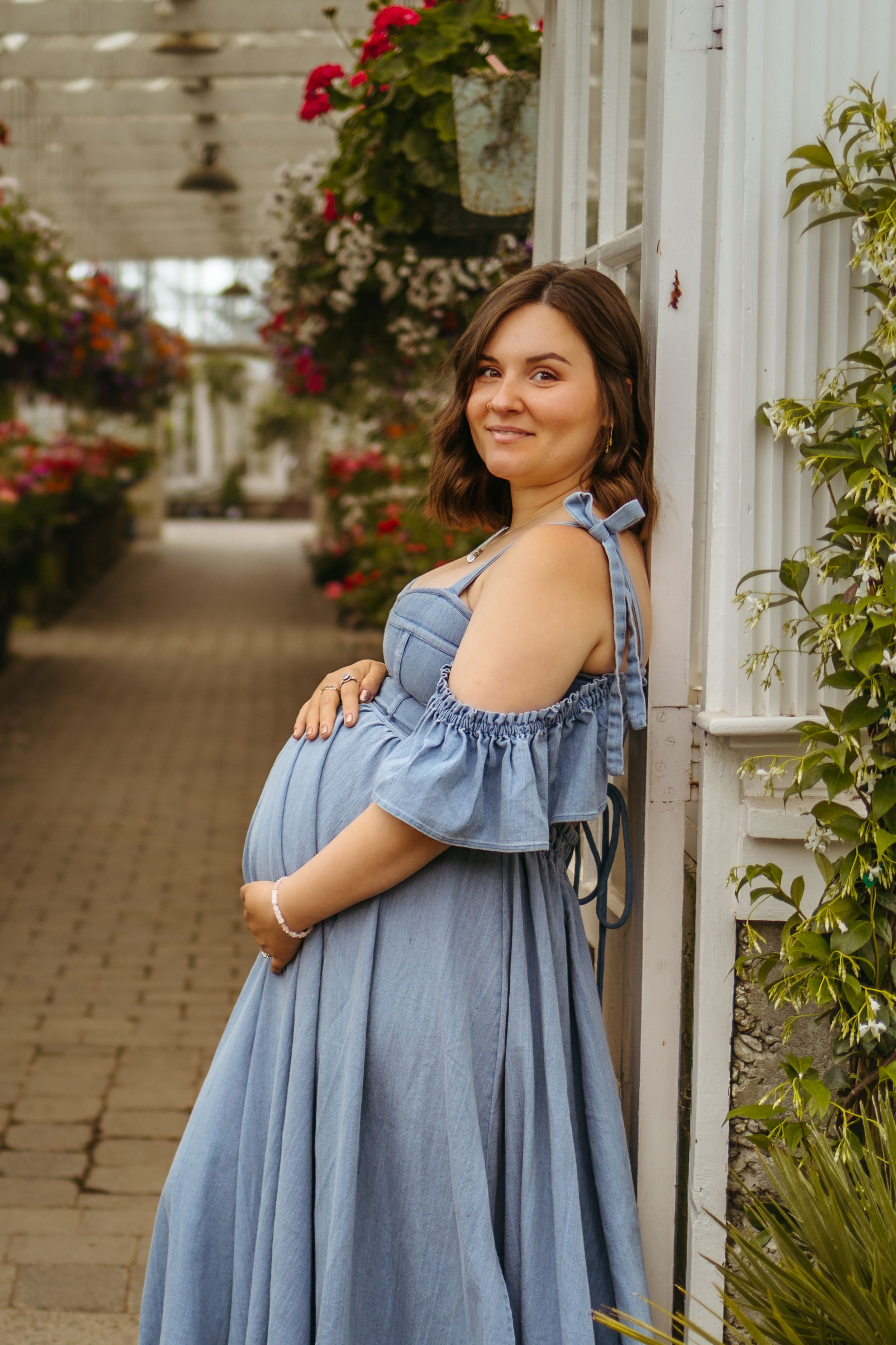 A person in a light blue ruffled dress poses gracefully against a white column with red flowers in the background.