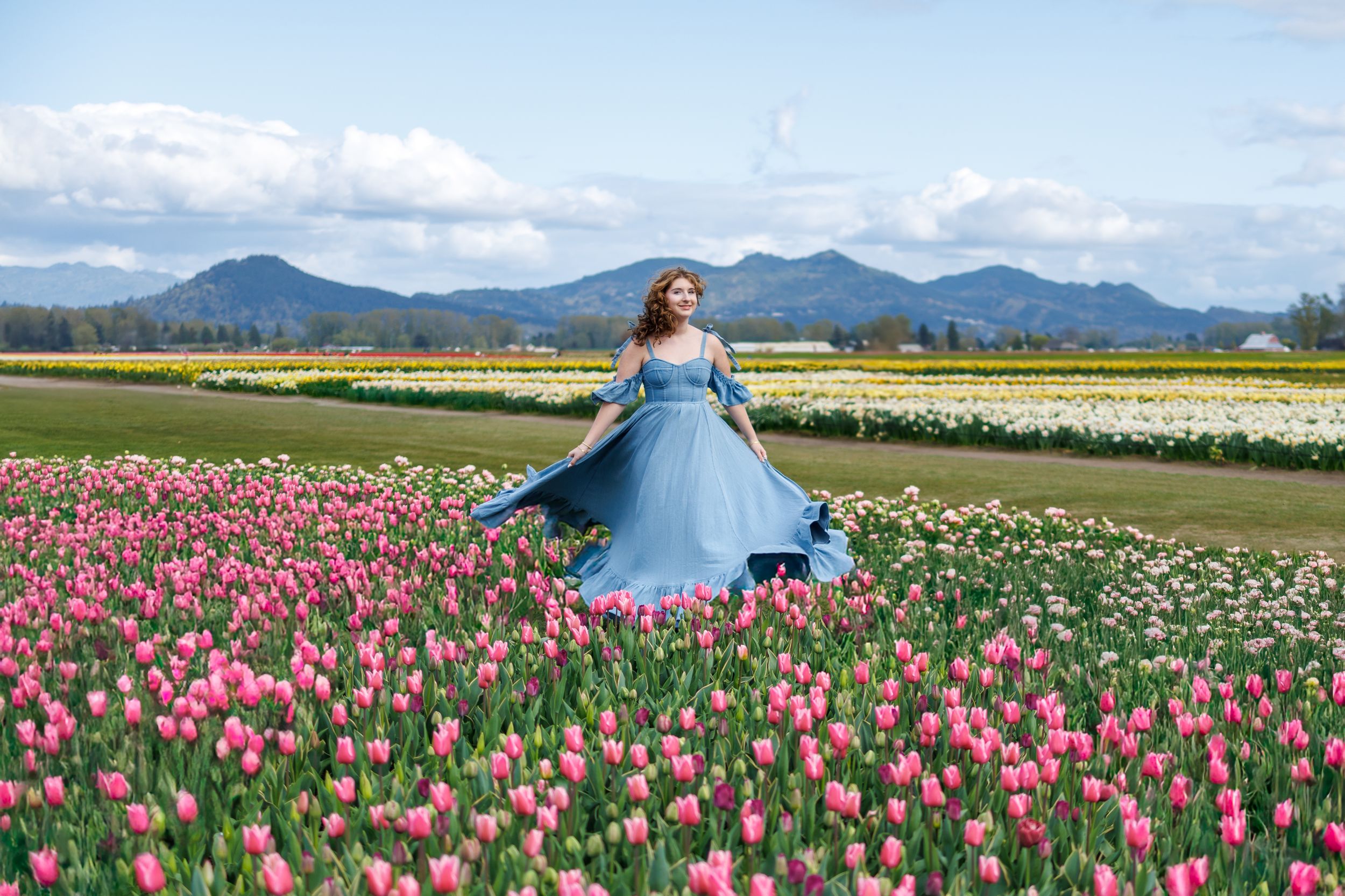 A person in a flowing blue dress twirls among vibrant pink tulip fields with mountains in the background.