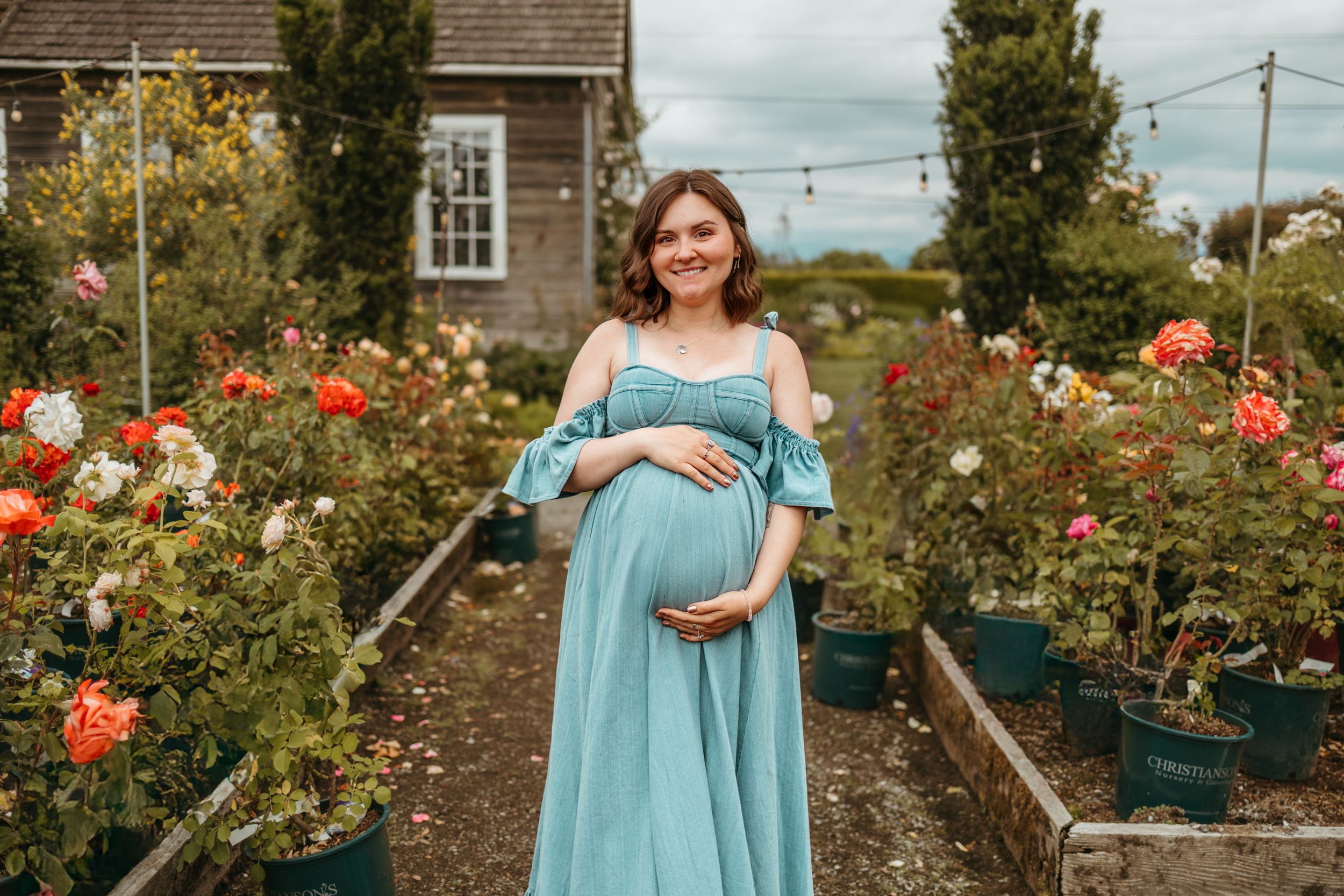 A woman in a turquoise dress stands in a blooming rose garden with red flowers and greenery surrounding her.