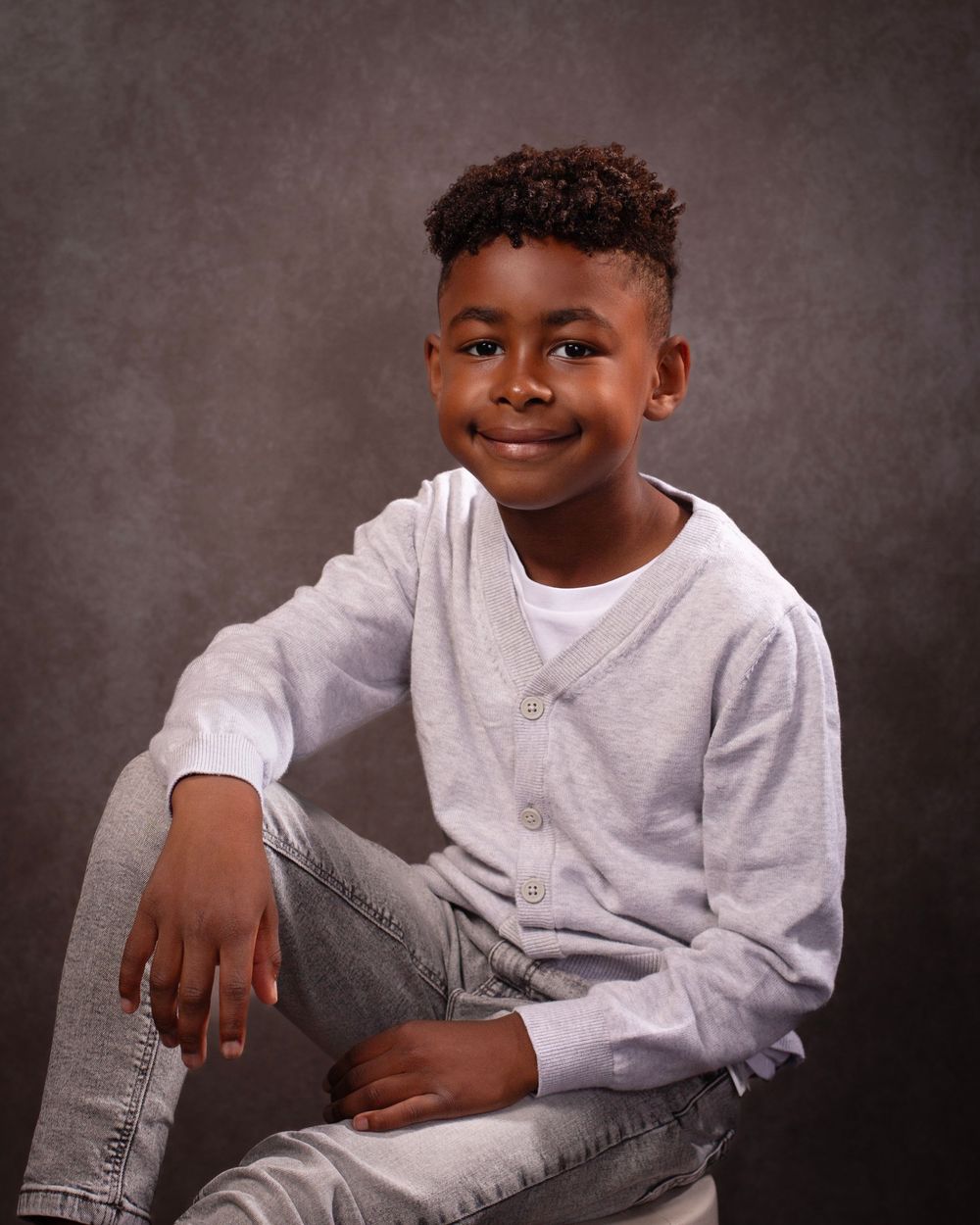 Young child poses confidently in a gray sweatshirt and jeans against a textured dark background.