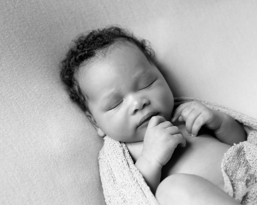 A peaceful sleeping newborn baby rests with thumb near mouth in black and white photograph.