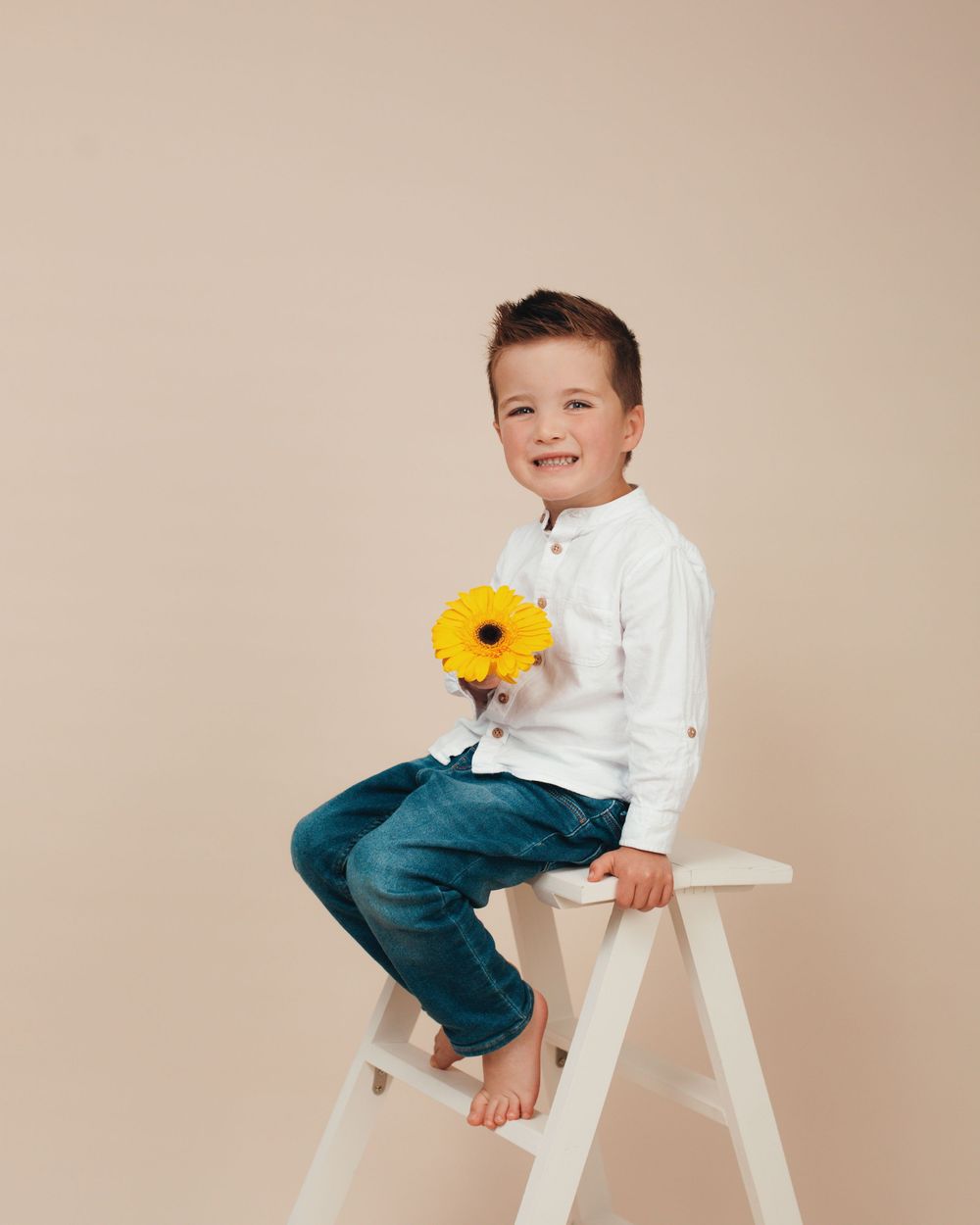 Child sitting on a white ladder stool wearing teal pants and white top while holding a yellow sunflower in a studio setting.