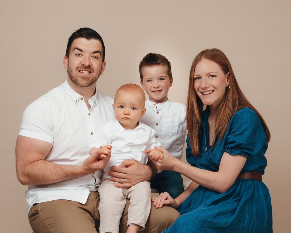 A family with an infant sits together on a beige couch, wearing coordinated white and blue outfits.