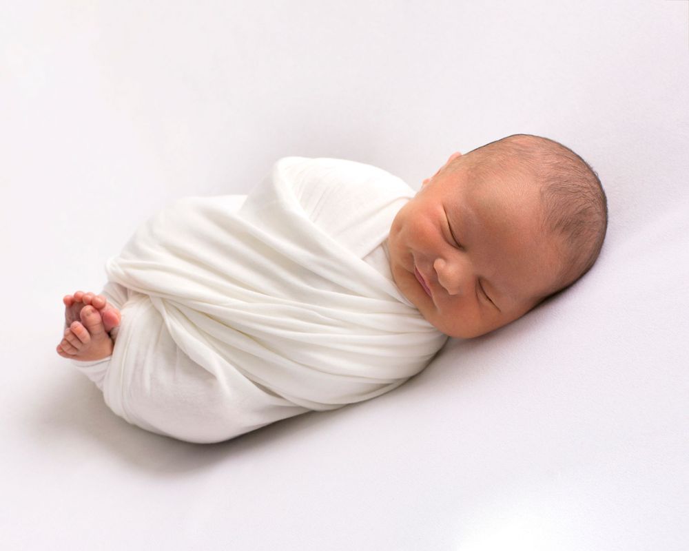 A newborn baby wrapped in white cloth photographed from multiple angles lying peacefully against a light background.