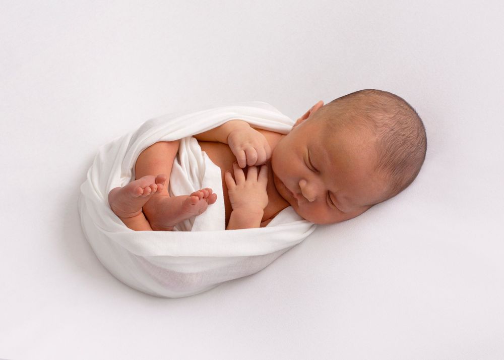 Sleepy newborn baby swaddled in white blankets lying on white background.