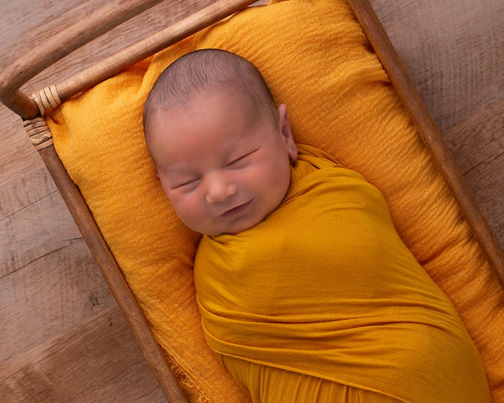 Sleeping newborn wrapped in bright yellow swaddling fabric rests peacefully on matching yellow cushion.