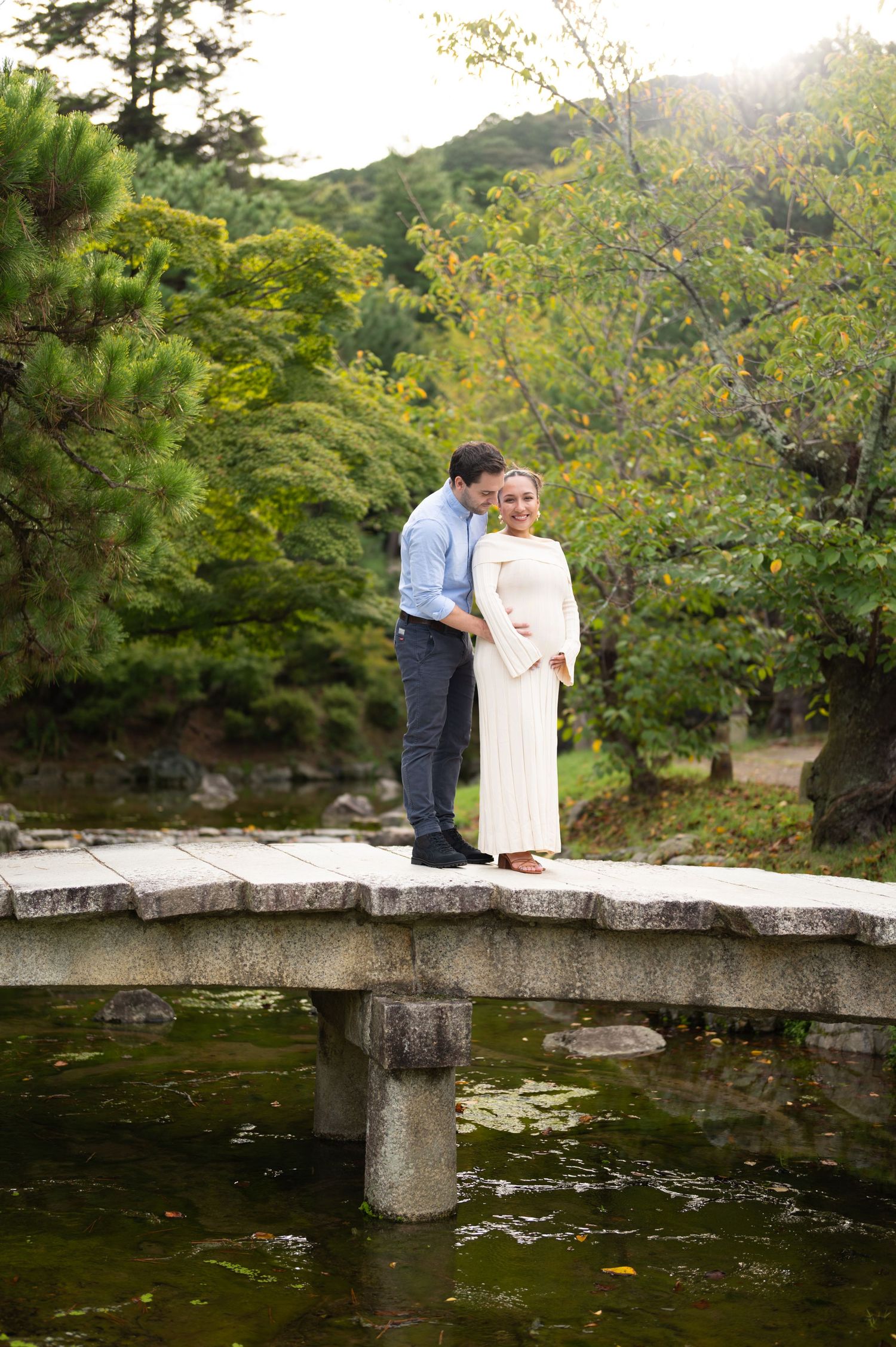 Couple embraces on a stone bridge over a pond in a tranquil Japanese garden at golden hour.