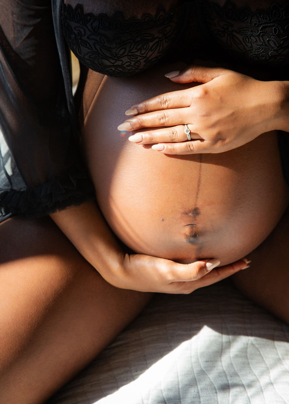 Close-up of a pregnant Black woman cradling her bare baby bump, wearing black lace lingerie and a diamond ring.