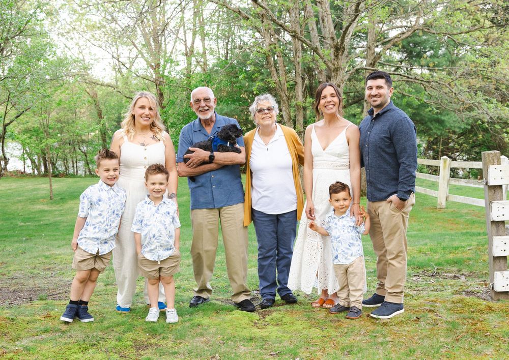 Multi-generational family group posing outdoors on green lawn with white fence, including children and adults holding a black dog.