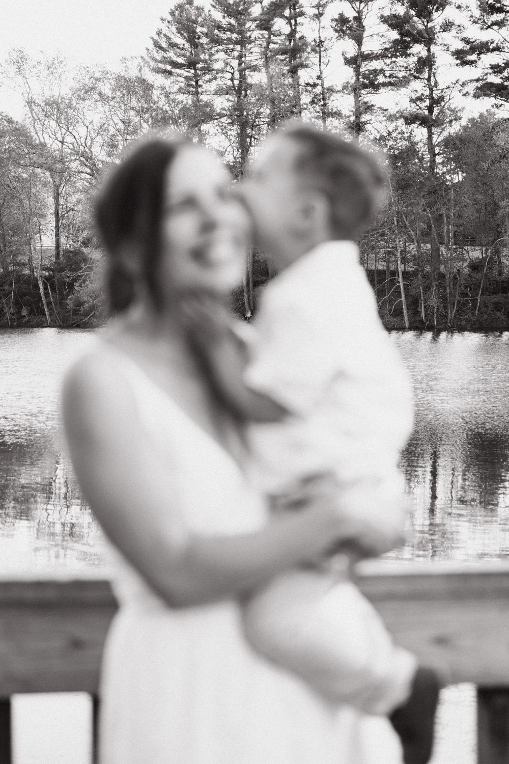 Black and white photo of a smiling woman in a white dress holding a baby near a serene lake with trees in the background.