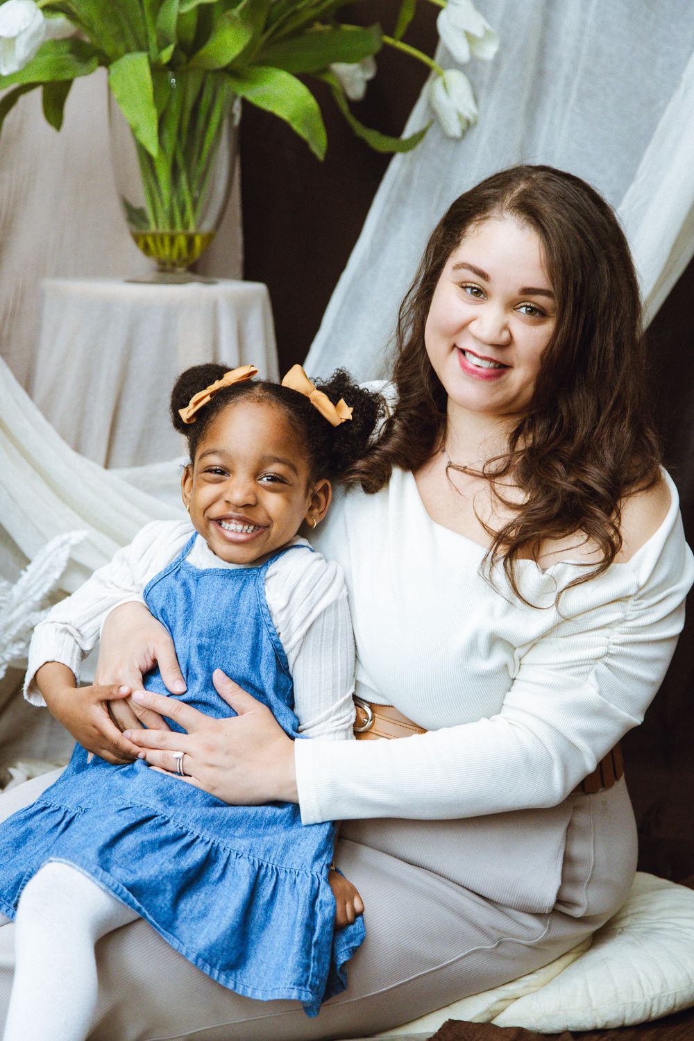 A woman smiling and hugging a young girl in a blue dress, sitting together in a cozy indoor setting with white tulips.