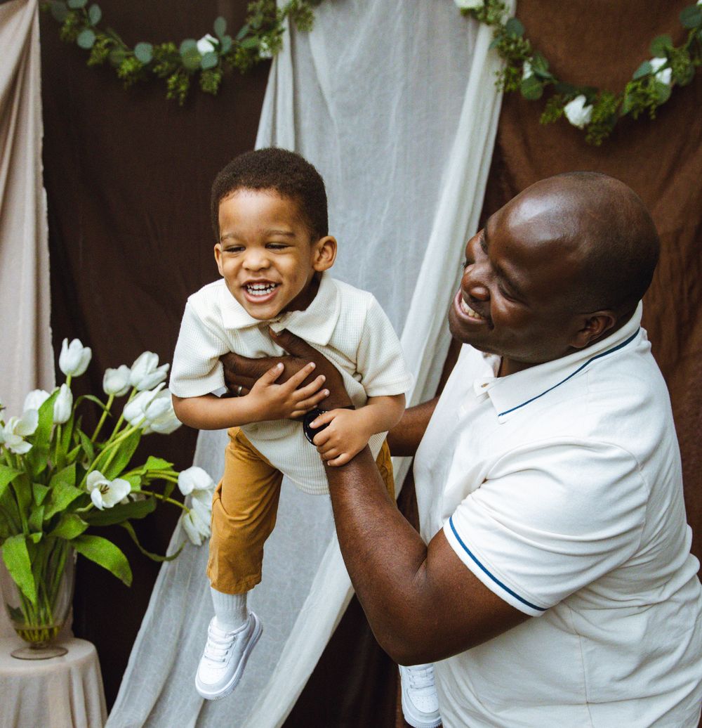 A joyful Black father lifts his laughing toddler son during a cheerful indoor photo session with white tulips.