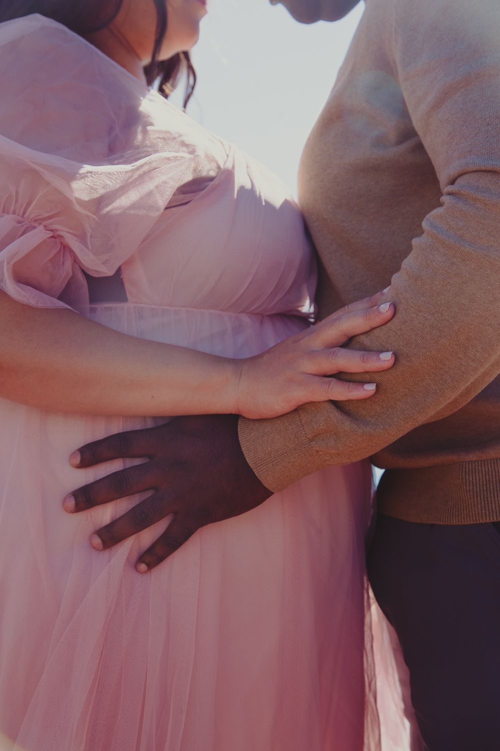 Interracial couple tenderly embracing pregnant belly, woman in pink tulle dress, maternity photo shoot.