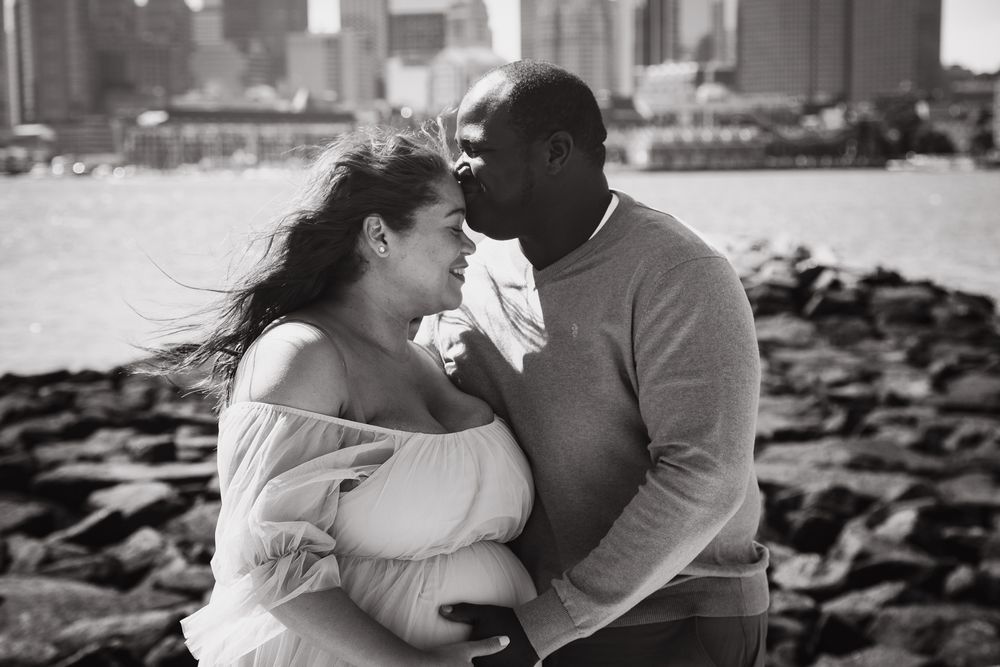 Black and white photo of a pregnant couple embracing near rocky waterfront with city skyline in background.