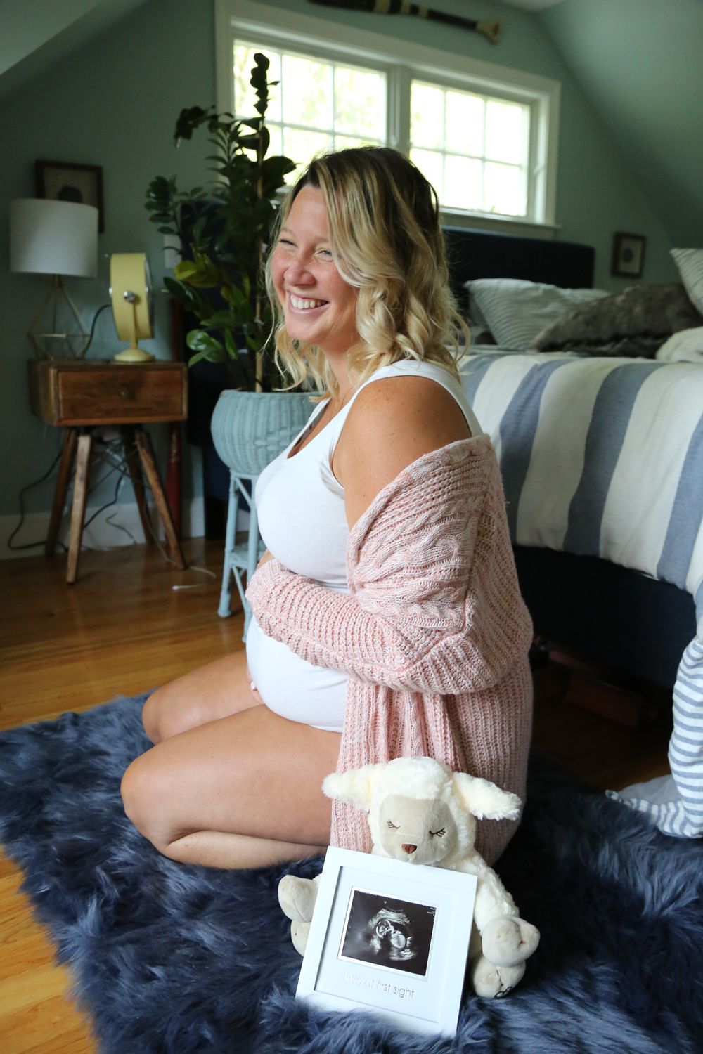 Smiling pregnant woman in pink cardigan sitting on blue fur rug holding baby ultrasound photo and stuffed animal toy.