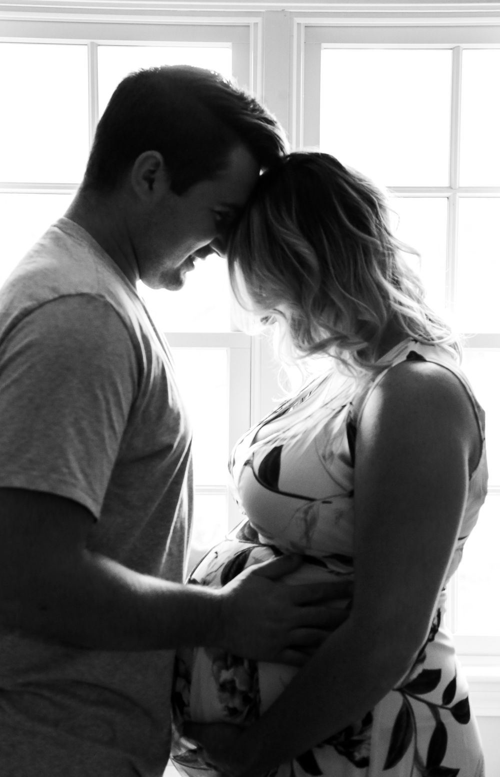 Black and white photo of a couple sharing an intimate moment, with the woman cradling a baby bump by a bright window.