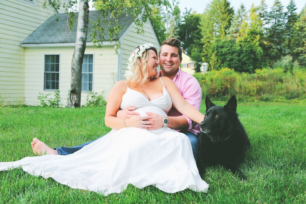 Pregnant woman in white dress sits with smiling man and black dog on green lawn near white cottage.