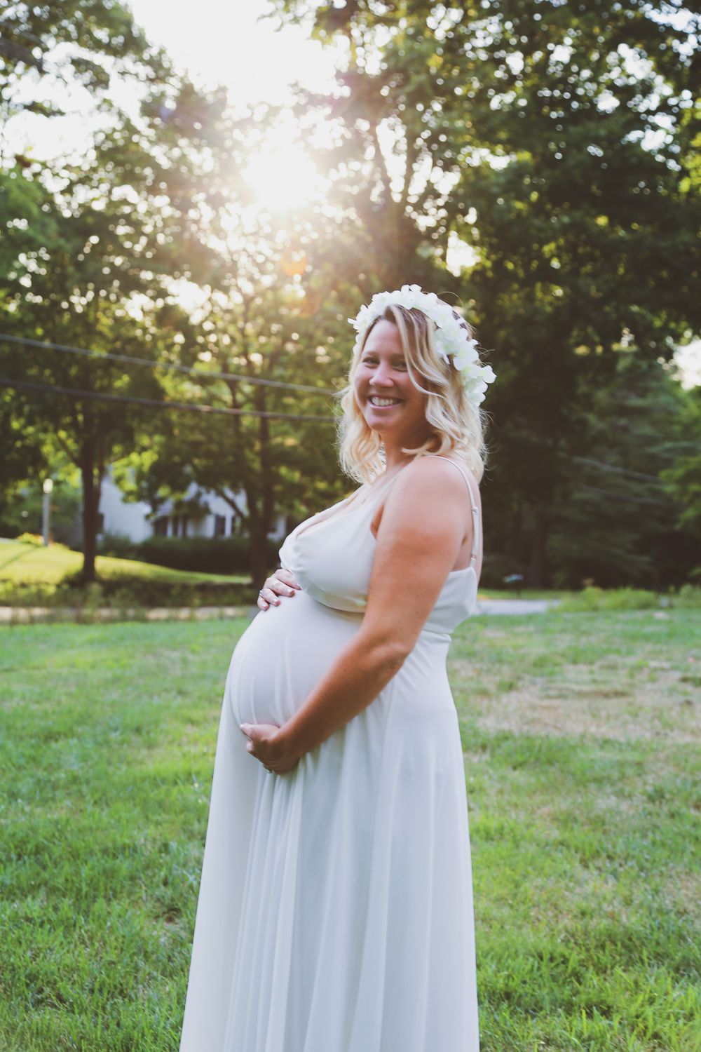 Smiling pregnant woman in white dress and floral crown holds baby bump outdoors at golden hour.