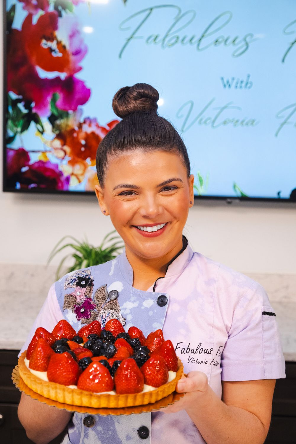 Chef in white uniform presents a beautiful fruit tart topped with fresh strawberries and berries against floral backdrop.