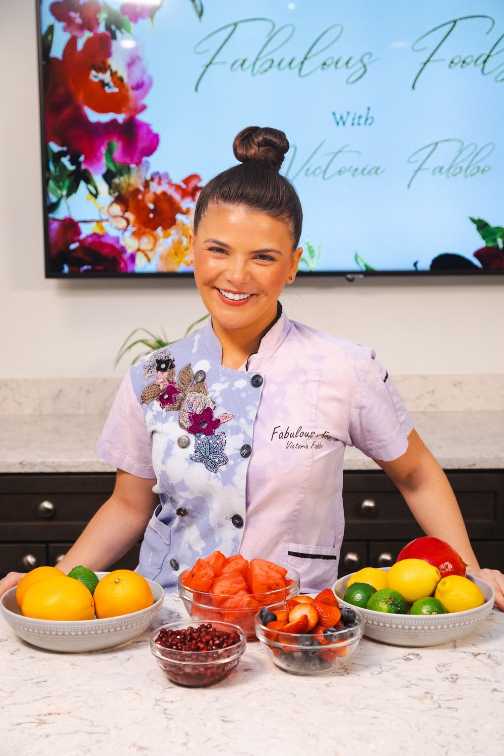 A cooking show setup displays colorful fresh fruits and ingredients in gray bowls on a white marble counter.