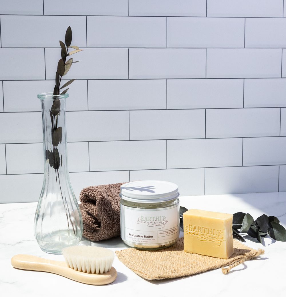 Natural bath and body care products arranged on white tile with wooden accessories and dried flowers.