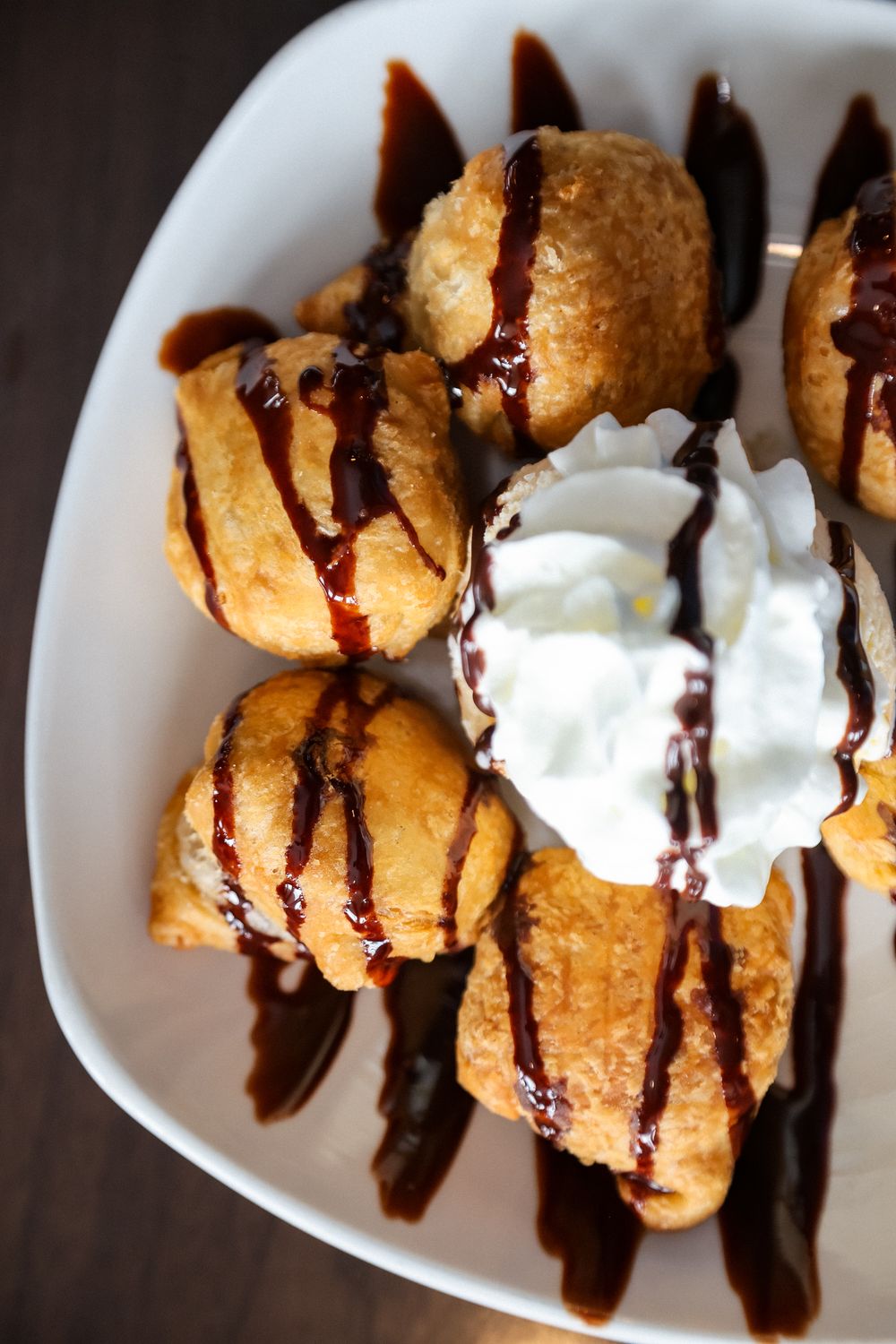 Golden cream puffs drizzled with chocolate sauce served on a white plate with whipped cream.