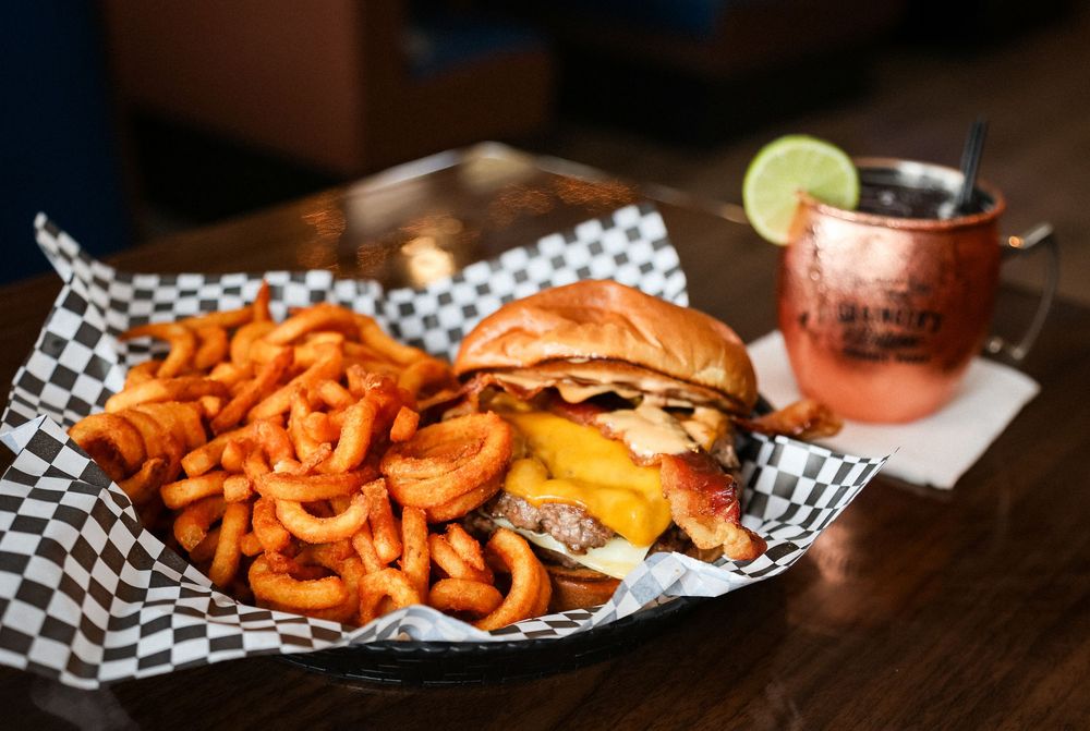 A juicy cheeseburger with curly fries in a checkered basket beside a Moscow Mule cocktail on a wooden table.