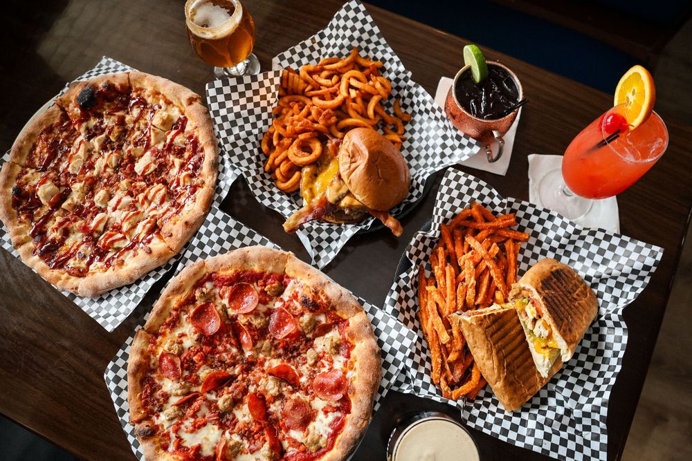 Overhead view of a restaurant spread featuring two pizzas, curly fries, sweet potato fries, a sandwich, and colorful drinks.