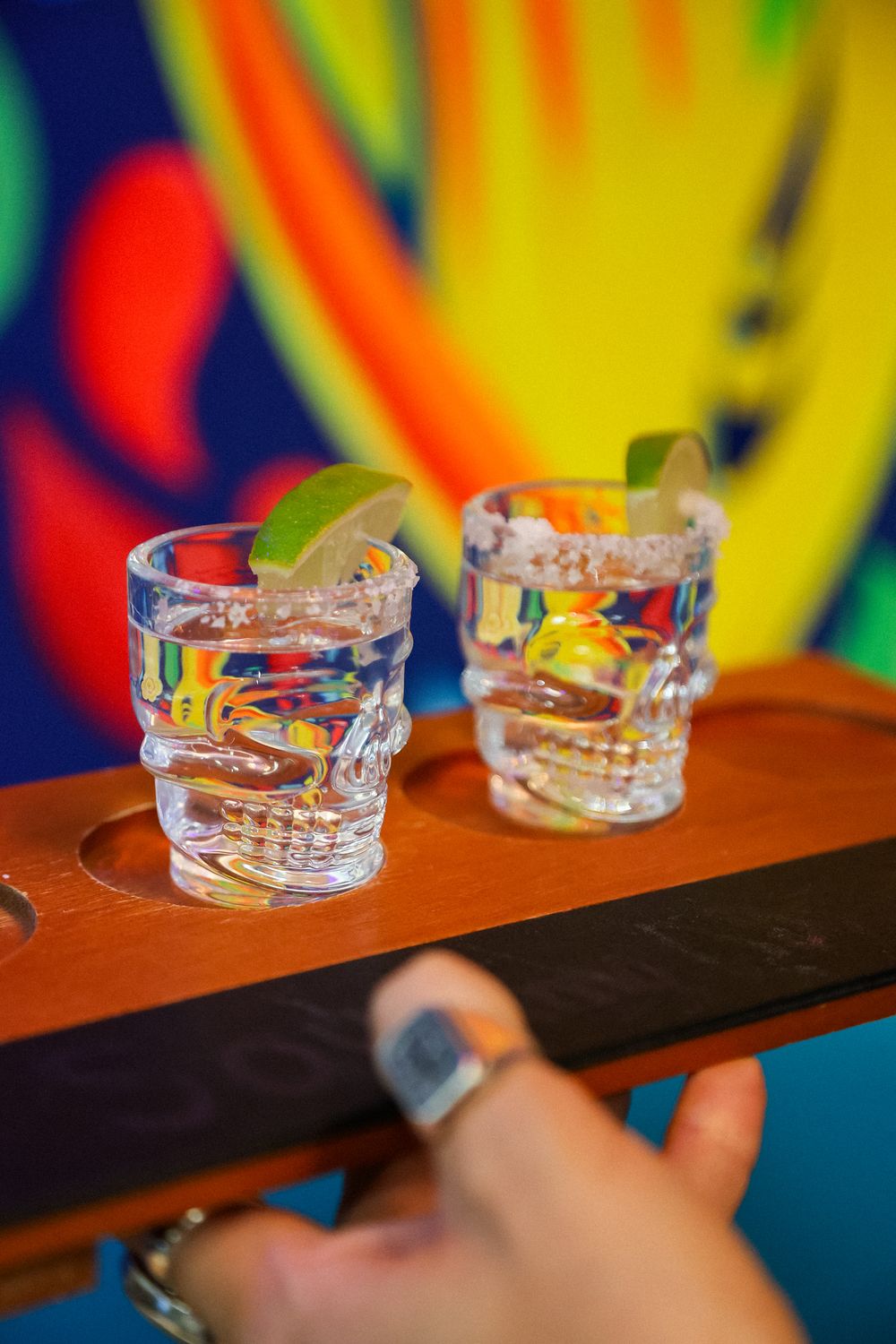 Two skull-shaped shot glasses with lime garnish on wooden tray, colorful mural background.