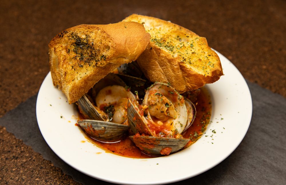 Steamed clams in tomato broth served in a white bowl with toasted garlic bread on top.