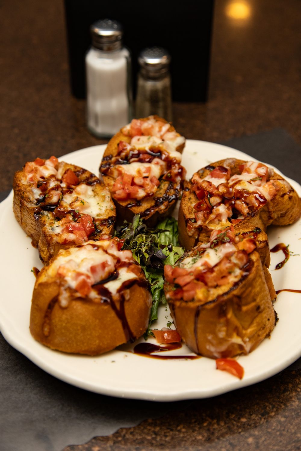 Bruschetta topped with tomatoes, melted cheese, and balsamic glaze served on toasted bread slices on a white plate.