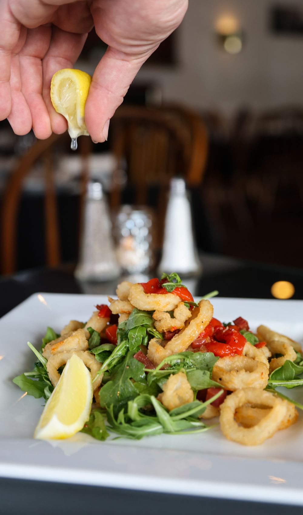Crispy fried calamari on arugula with roasted peppers, lemon wedge, and a hand squeezing fresh lemon juice over the dish.
