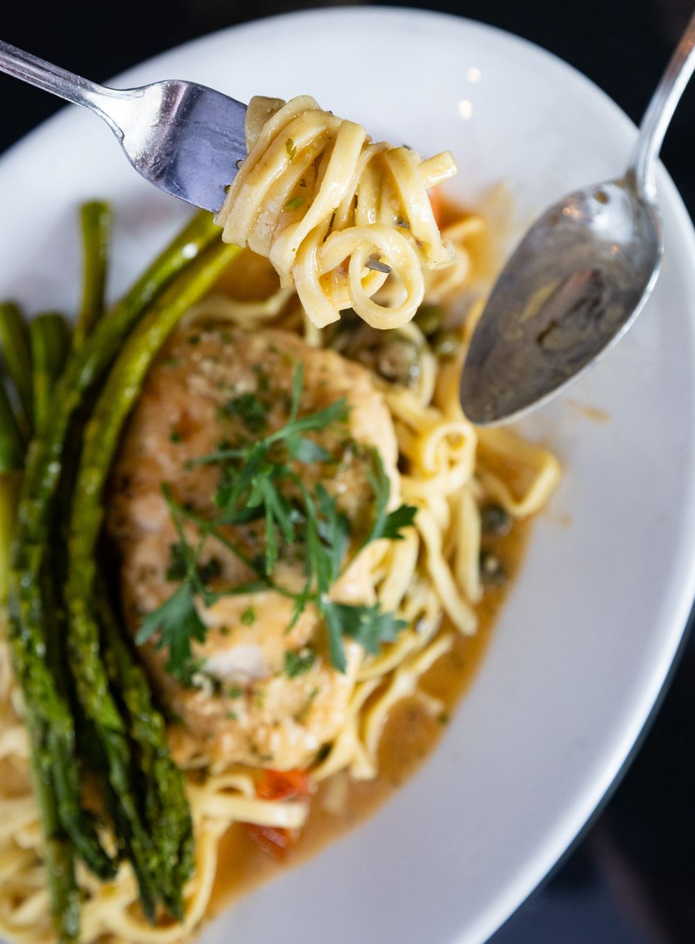 Fork twirling linguine pasta with shrimp, capers, and asparagus in a savory broth on a white plate.