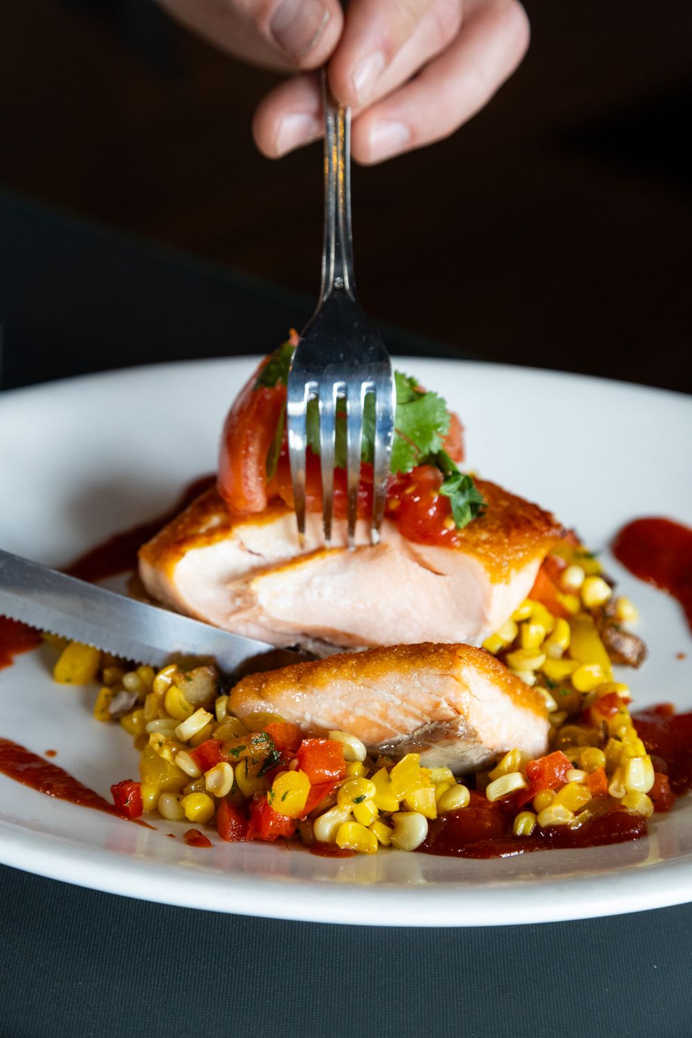 Hand cutting into a seared salmon fillet served over corn succotash with red pepper sauce on a white plate.