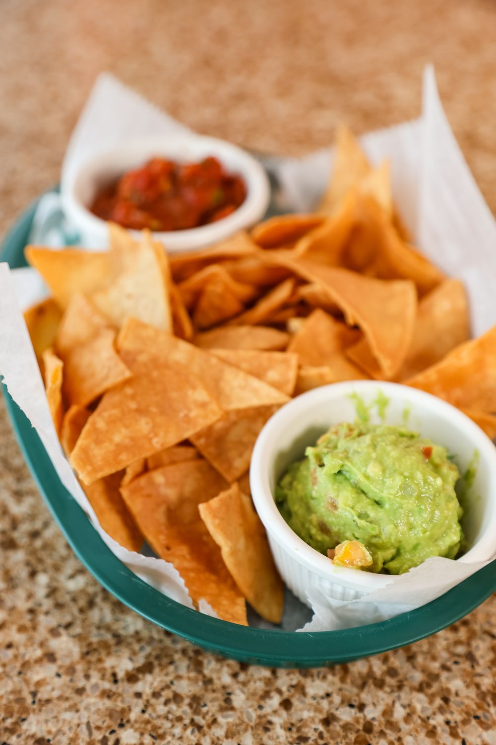 Tortilla chips served in a basket with guacamole and salsa dipping sauces on a granite countertop.