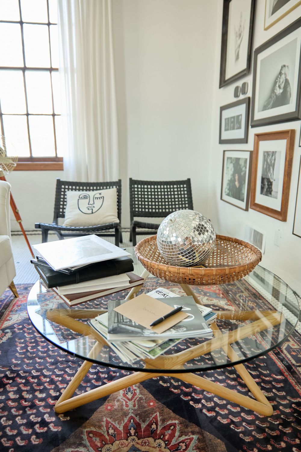 Glass coffee table with wooden base surrounded by black chairs on a patterned area rug.